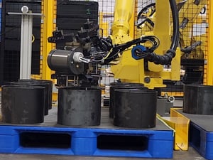 The image shows an industrial robotic arm equipped with a gripping and vision system hovering over several black cylindrical blocks on a blue pallet. It's an automated system on a manufacturing floor, designed for efficiency and precision in handling materials. A grid encloses it, a safety measure.